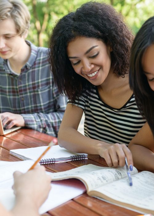 Image of multiethnic group of young happy students sitting and studying outdoors while talking. Looking aside.